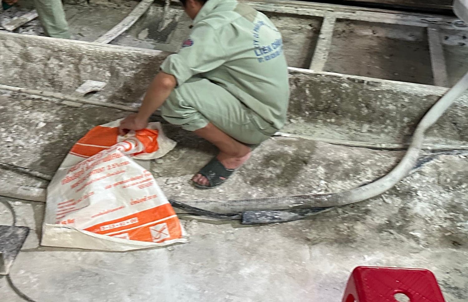 A factory floor drainage channel opened for cleaning. A worker holds a woven collection sack while lime deposit is cleared; a hose runs across concrete caked with white scale.