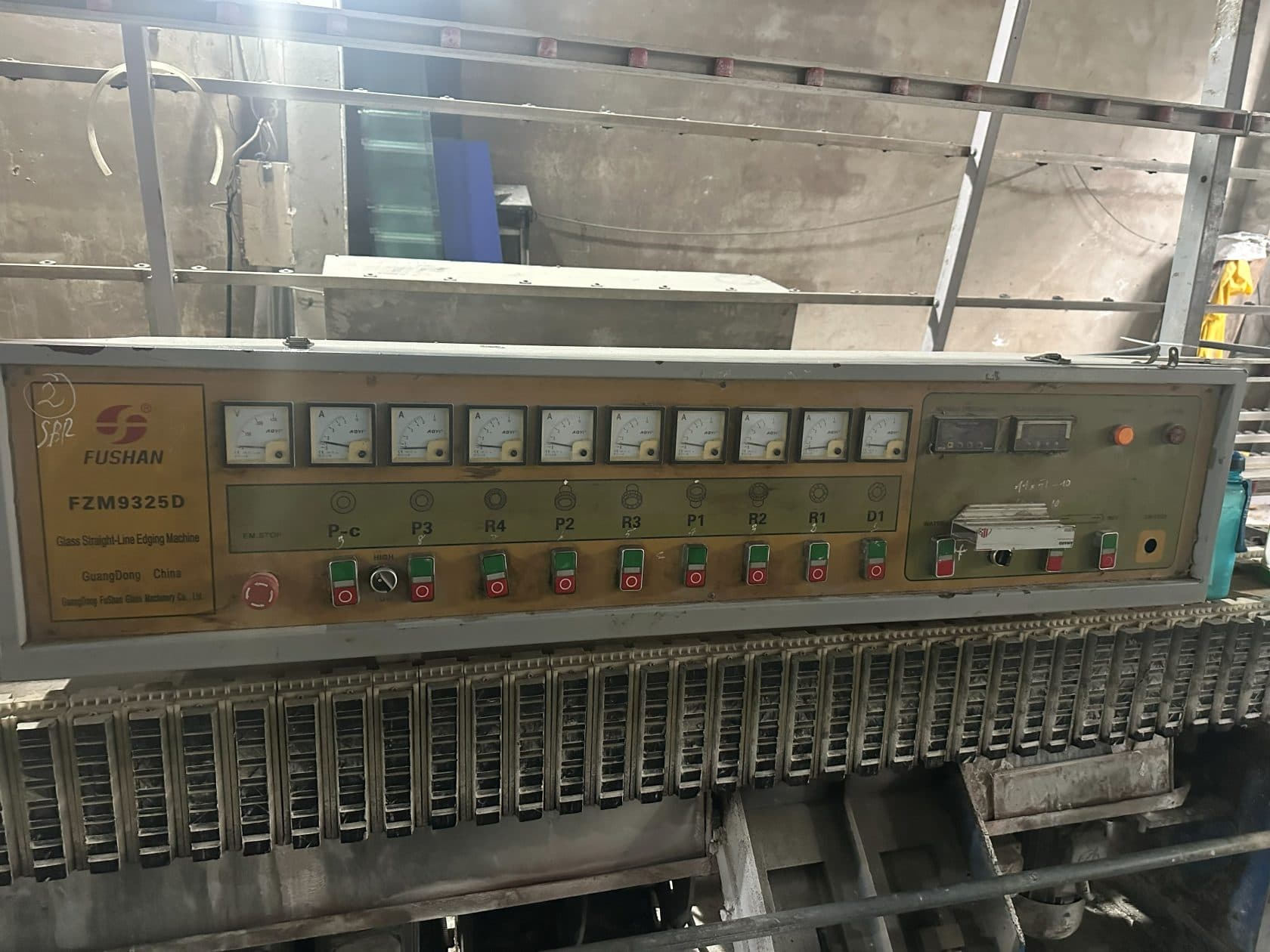 The control panel of a Fushan FZM9325D straight-line glass edging machine. A row of nine analogue ammeters sits above nine labelled station controls, marked from right to left D1, R1, R2, P1, R3, P2, R4, P3 and P-c, each with a green and a red button. The panel is worn and dusted with grinding residue; the conveyor chain of the machine runs below it.